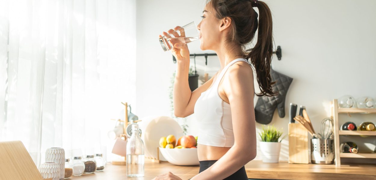 Asian beautiful woman in sportswear drink water after exercise at home. Young thirsty active sport girl takes a sips of clean mineral natural in cup after workout for health care in kitchen in house.